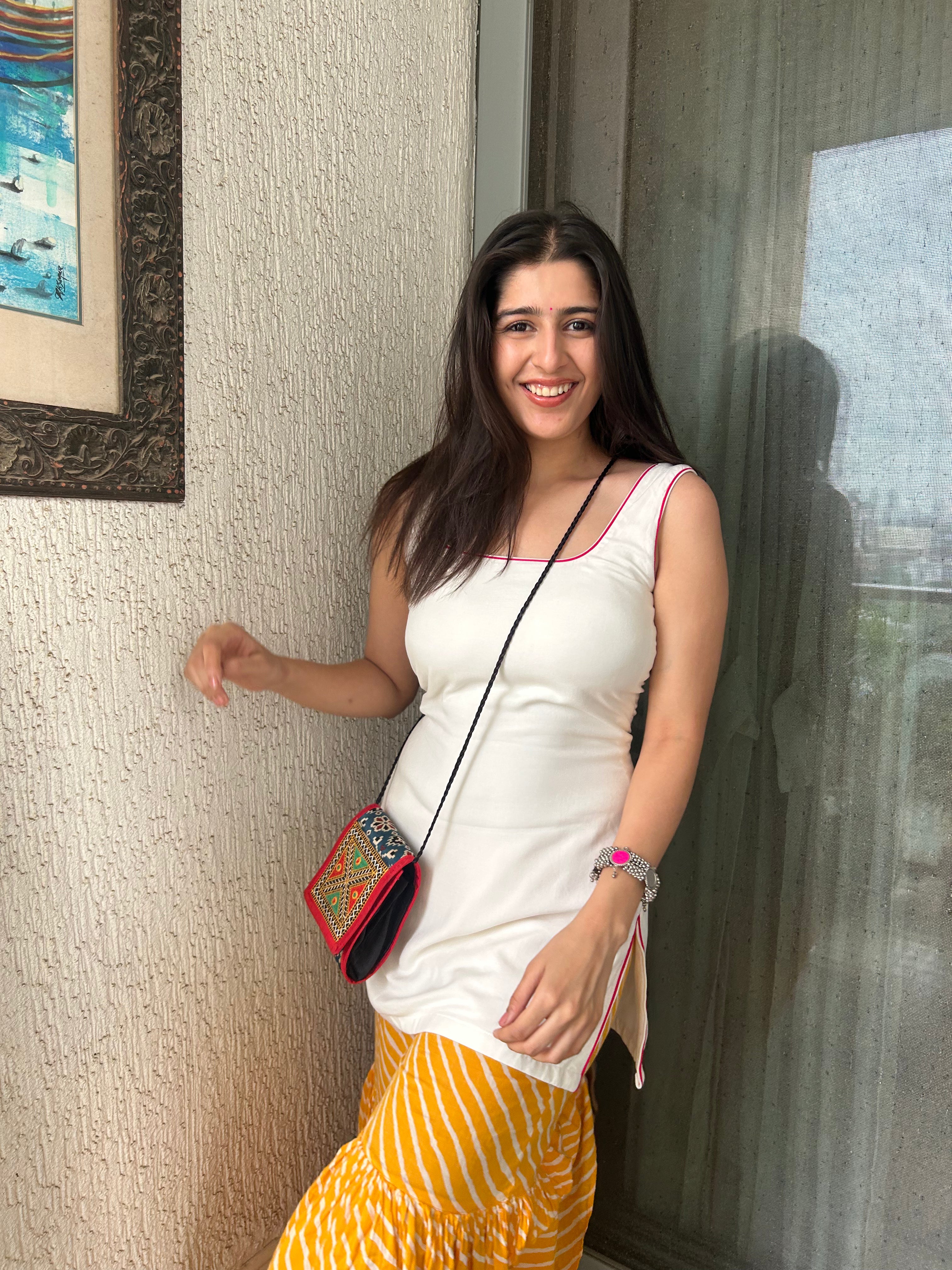 Aditi Singh in a Bindee Basics white tank-top Kurti and yellow skirt standing against a textured wall with a painting in the background.