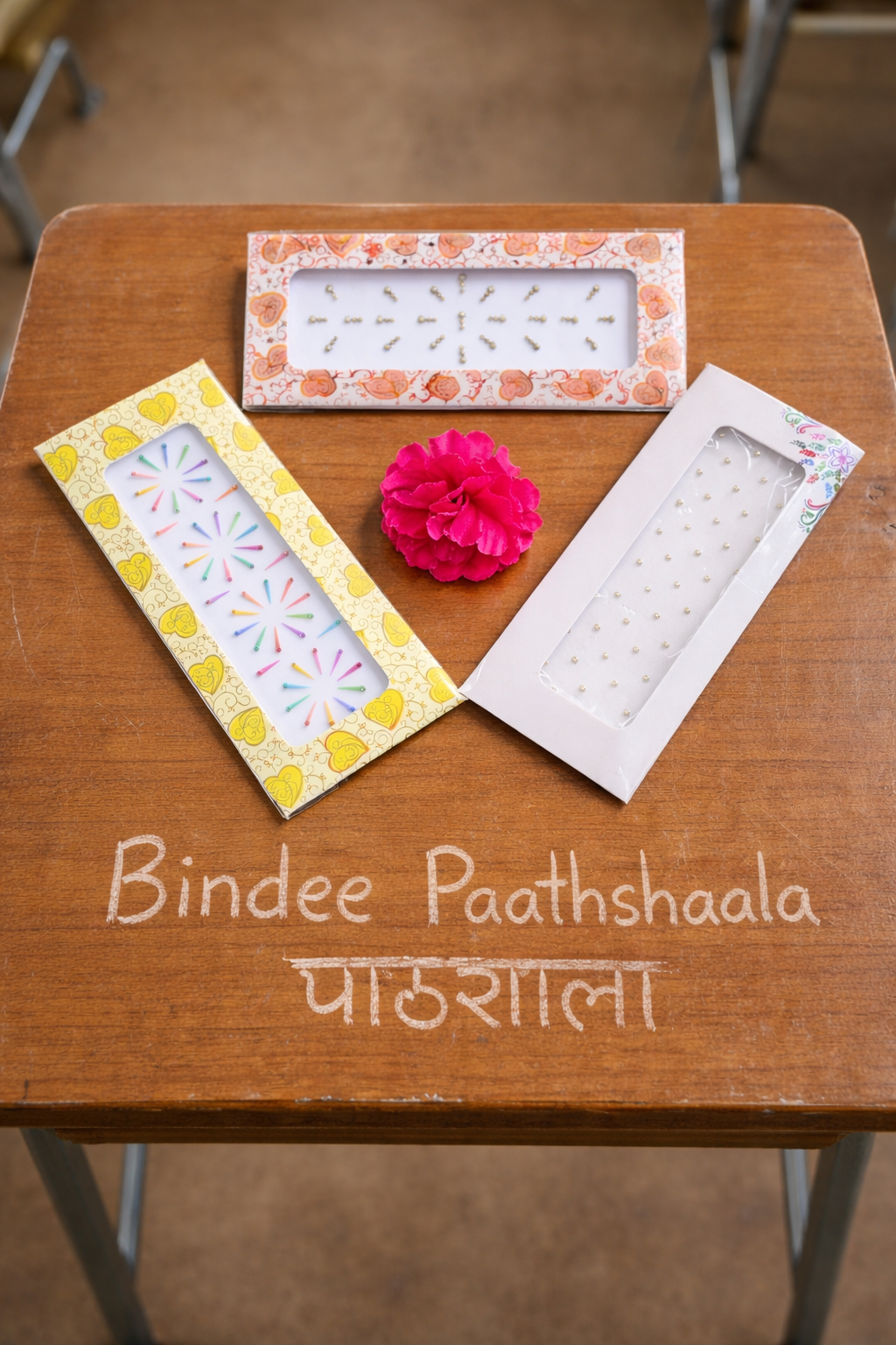 Three decorative Bindi packets on a wooden surface forming a downward triangle, with a pink flower and text 'Bindee Paathshaala'.
